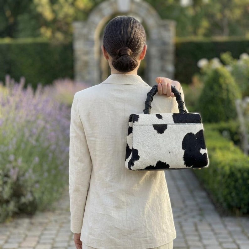 May include: A structured handbag featuring a black and white cow print pattern with a textured finish. The bag has a single top handle with a braided design and a rectangular silhouette. It is paired with a light beige blazer worn over the shoulders.