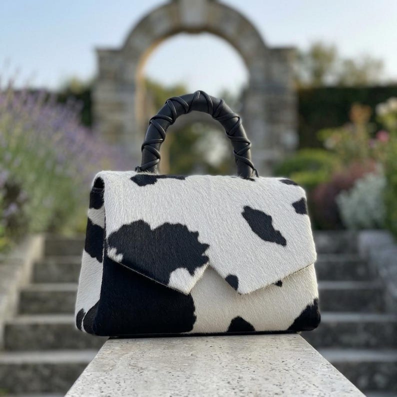 May include: A structured handbag featuring a black and white cow print pattern on a textured hide material. The bag has a flap closure and a single top handle wrapped in black leather, providing a distinct contrast against the spotted body.