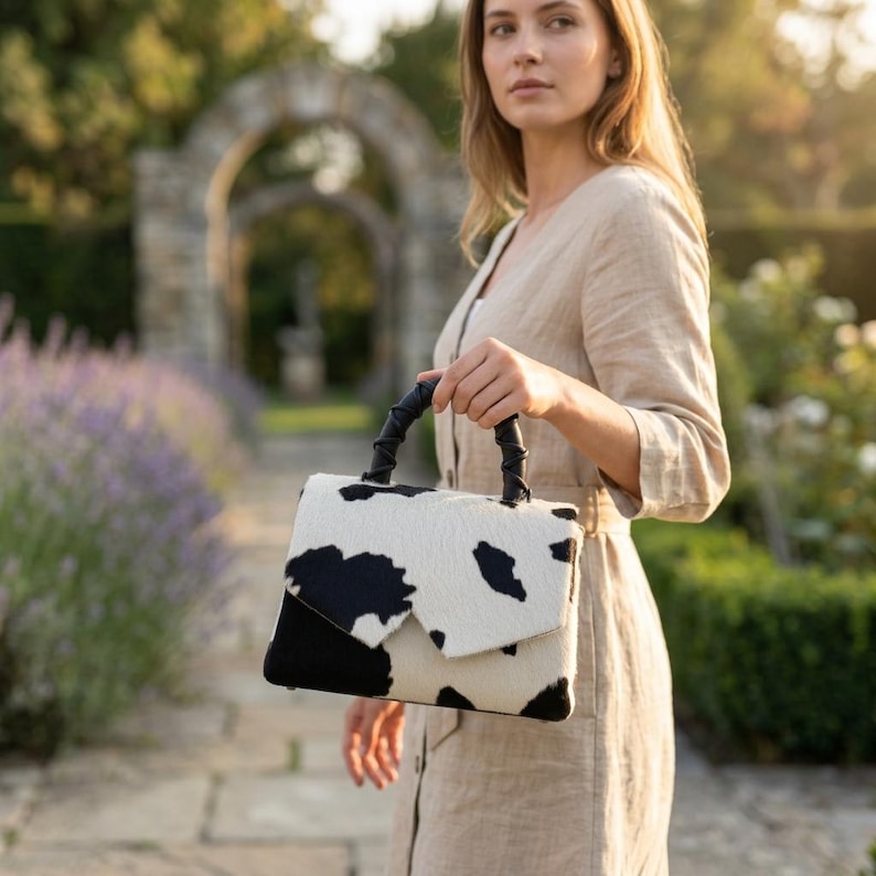 May include: A structured handbag featuring a black and white cow print pattern on a textured surface. The bag has a single top handle wrapped in black material and a flap closure that secures the main compartment with a clean, geometric design.