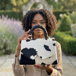 May include: A structured handbag featuring a black and white cow print pattern with a textured hair-on-hide finish. The bag has a flap closure and a black leather-wrapped top handle with a twisted design, providing a distinct visual contrast.