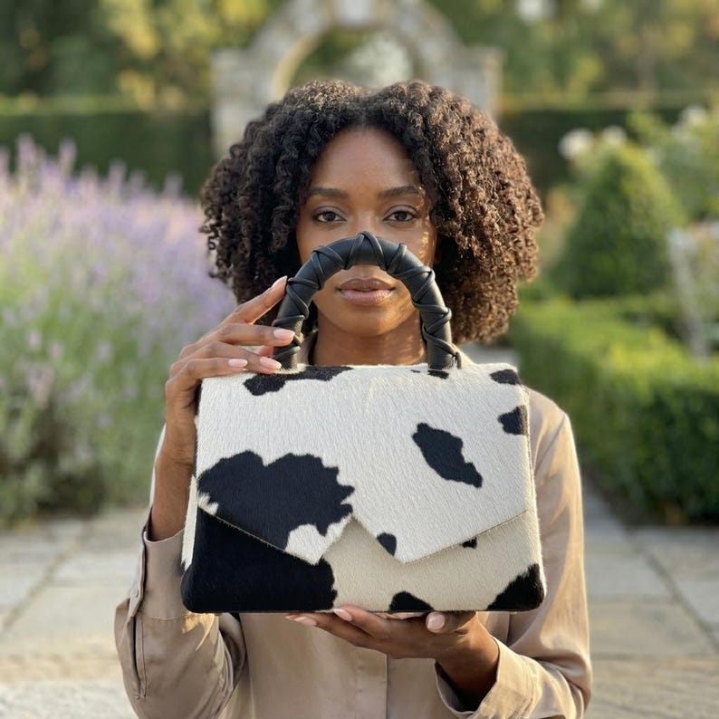 May include: A structured handbag featuring a black and white cow print pattern with a textured hair-on-hide finish. The bag has a flap closure and a black leather-wrapped top handle with a twisted design, providing a distinct visual contrast.