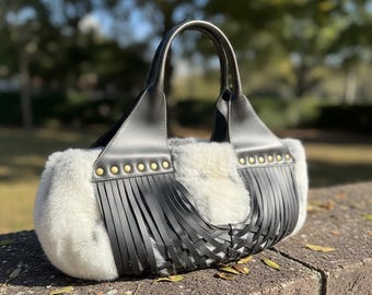 May include: A black and white handbag with a soft, fluffy texture and leather accents. The bag features a woven design, fringe detailing, and gold-colored stud embellishments. The handles are black and the bag is sitting on a brick wall.