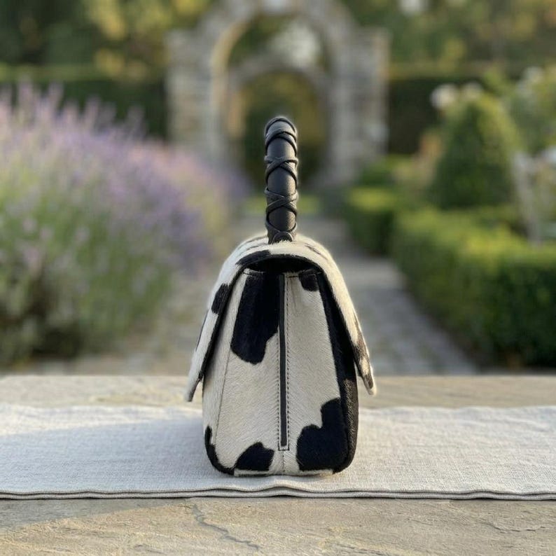 May include: A structured handbag featuring a black and white cow-print pattern with a textured hair-on-hide finish. The bag includes a single top handle with a braided black leather design and a flap closure that secures the main compartment.