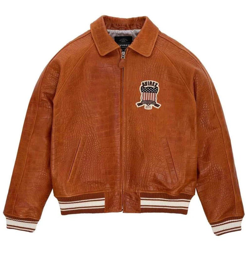 May include: A brown leather jacket with a crocodile texture. The jacket has a zipper closure, a collar, and a patch on the chest with the text "AVIREX". The cuffs and hem have white and brown stripes.