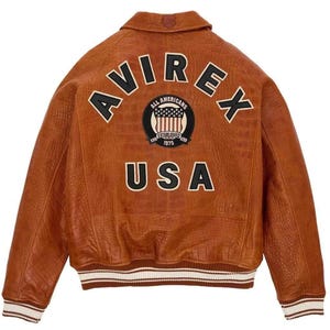 May include: A brown leather jacket featuring the word "AVIREX" in white lettering across the back. Below is a patch with "ALL AMERICANS" and "ESTABLISHED 1975". The word "USA" is below the patch. The jacket has a ribbed white and brown trim.