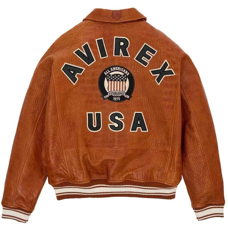 May include: A brown leather jacket featuring the word "AVIREX" in white lettering across the back. Below is a patch with "ALL AMERICANS" and "ESTABLISHED 1975". The word "USA" is below the patch. The jacket has a ribbed white and brown trim.