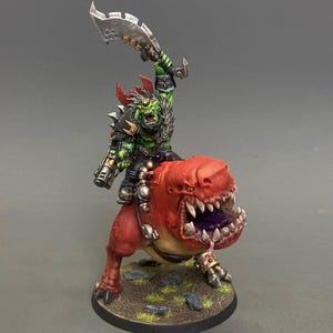 Puede incluir: Figura en miniatura de un orco de piel verde montado sobre una criatura roja con aspecto de dinosaurio. El orco sostiene una gran cuchilla dentada. La criatura tiene una boca abierta con dientes afilados. La base es circular y está pintada para parecer una zona herbosa.
