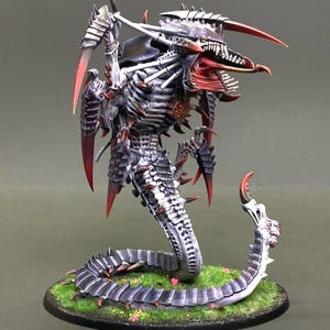 May include: A detailed miniature figure of a Tyranid creature, featuring a segmented body, sharp claws, and a menacing head. The figure is painted in shades of gray, red, and black, and is posed on a base with green grass and small flowers.