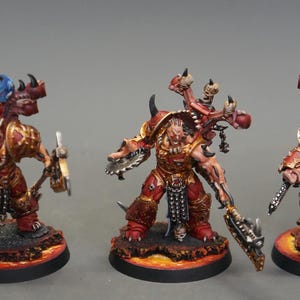 May include: Three miniature figurines of armored warriors, each with a red and gold color scheme. The figures have detailed features, including weapons and grotesque facial expressions. They stand on circular bases with a fiery design, suggesting a fantasy or sci-fi theme.