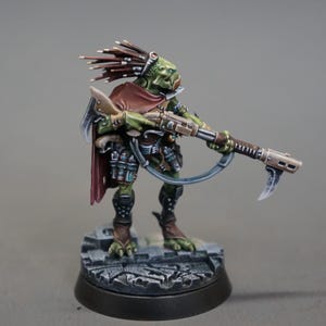 May include: A miniature figurine of a green-skinned alien warrior, holding a rifle with a curved blade attachment. The figure wears a red cape and has a headdress of brown spikes. It stands on a textured gray base.