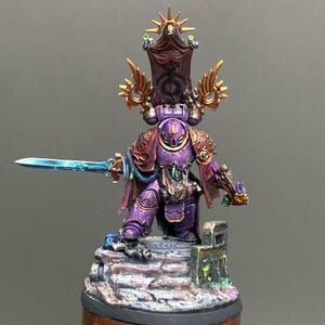 May include: A miniature figurine of a warrior in purple armor, holding a glowing blue sword. The figure has gold accents, a red cape, and a banner above its head. It stands on a stone base with green and blue details.