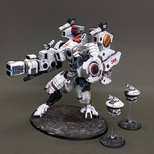May include: A detailed miniature model of a white and gray mech with red and black accents. The model features a large cannon, multiple missile launchers, and two smaller drone-like units. The mech is posed on a textured black base.