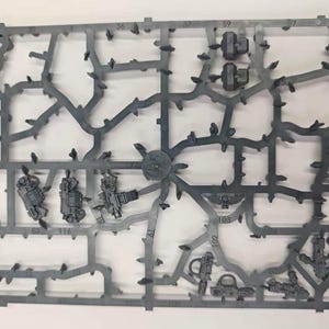 May include: A gray plastic model kit sprue featuring various unattached miniature components, including weapon parts, small mechanical accessories, and an emblem with a skull, all connected by a grid-like frame with numbered identification tags.