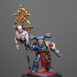 May include: A miniature figurine of a blue and gold armored warrior holding a staff with a gold emblem and a chained, pale-skinned figure. The warrior has red accents and is posed on a red base.