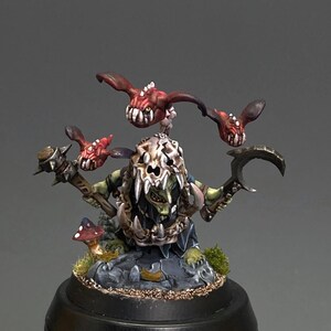 May include: A miniature goblin figure wearing a large skull helmet, holding a spiked mace and a curved blade. Three small, winged red creatures with sharp teeth hover above the goblin. A small mushroom sits on the rocky base below.