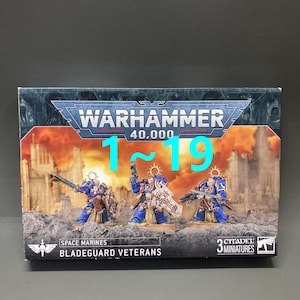 以下が含まれることがあります： ウォーハンマー40,000のミニチュアのボックスセット。3体のスペースマリーン・ブレードガード・ベテランズが含まれています。ボックスにはウォーハンマー40,000のロゴと1〜19の数字がターコイズ色で表示されています。ミニチュアは青と金で、戦場をイメージしたベースに配置されています。