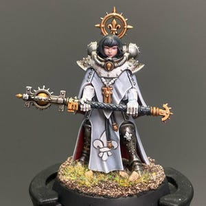 Op de afbeelding: Miniatuurfiguur van een vrouw in een wit en zilver uniform, met een staf. De figuur heeft zwart haar en een halo. De basis is bedekt met gras. De figuur is een miniatuur van een personage uit een sciencefiction- of fantasiespel.
