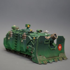 May include: A detailed miniature model of a green military vehicle with a large cannon and a front blade. The model features intricate details, including tracks, hatches, and a red cross emblem. The vehicle is painted in shades of green and brown.