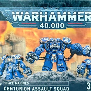 Op de afbeelding: Een Warhammer 40,000 Space Marines Centurion Assault Squad doos met drie blauw gepantserde miniatuurfiguren met gouden metalen details. De verpakking toont de productnaam en bevat in totaal 3 Citadel-miniaturen.