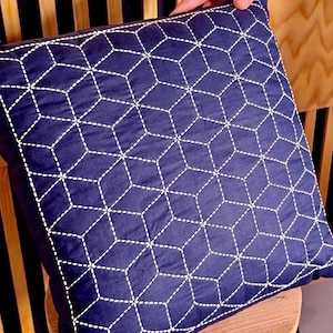 Pode incluir: Almofada quadrada azul marinho com um padrão geométrico de cubo costurado com fio branco. A almofada é feita de um tecido macio e provavelmente é usada para decoração de casa. A almofada tem aproximadamente 45 cm quadrados.