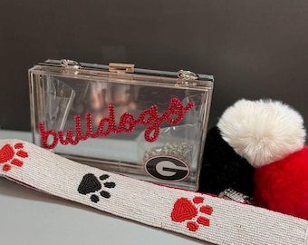 Puede incluir: Un embrague de acrílico transparente con la palabra "bulldogs" en escritura roja. Una correa de cuentas blancas con huellas de patas rojas y negras está al frente. Un llavero de pompón rojo, negro y blanco está a la derecha.