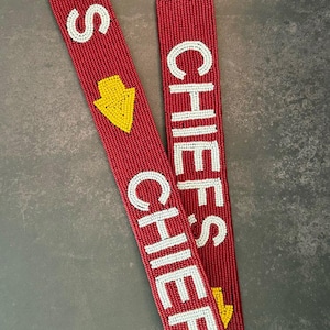 May include: Red beaded lanyard with the word "CHIEFS" in white beads. A yellow arrow points down. The lanyard has a gold-colored clasp.