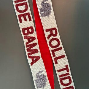 Pode incluir: Cinto de contas marfim com as palavras "ROLL TIDE BAMA" em letras carmesim. O cinto tem um verso de veludo vermelho. Dois desenhos de elefantes cinzentos também são apresentados no cinto.