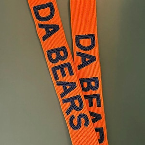Puede incluir: Cordón de cuentas naranja con el texto "DA BEARS" en azul marino. Tiene un cierre dorado. El diseño está hecho completamente de cuentas.