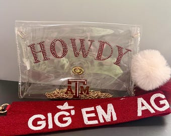 Könnte beinhalten: Eine durchsichtige Umhängetasche aus Kunststoff mit goldenem Drehverschluss und goldener Kette. Die Tasche zeigt das Wort HOWDY aus roten Strasssteinen und ein ATM-Logo. Sie enthält einen roten, perlenbesetzten Riemen mit weißem Text GIG EM AG und einen weißen Bommel.