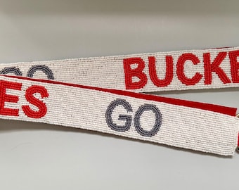 May include: A white beaded strap with the words "EYES GO BUCKEY" in red and gray beads. The strap has gold-colored metal clasps on the ends and a red fabric backing.