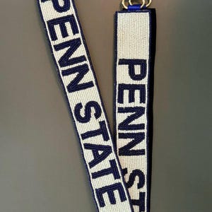 May include: A beaded lanyard with the text "PENN STATE" in navy blue letters on a white background. The lanyard has a gold-colored clasp and navy blue trim.