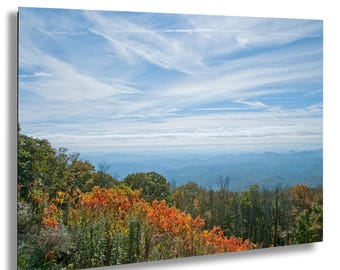 May include: A scenic landscape print featuring a mountain range under a blue sky with wispy clouds. The foreground showcases trees with vibrant autumn foliage in shades of red, orange, and green. The artwork captures a serene natural vista.