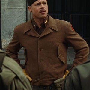 May include: A man in a brown military jacket with a dark green hat. The jacket has a button-up front, a collar, and a small pocket. The man is wearing a dark brown shirt underneath. The hat has a gold emblem.