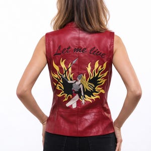 Può includere: Gilet in pelle rosso dal design senza maniche. La schiena presenta un disegno ricamato di una figura di guerriero con una lancia, circondata da fiamme gialle, e la scritta "Let me live" in nero.
