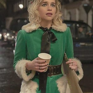 May include: A green velvet coat with faux fur trim and a black bow. The coat is belted at the waist with a brown belt. The person is holding a paper cup and a wooden sign, and wearing a floral crown.