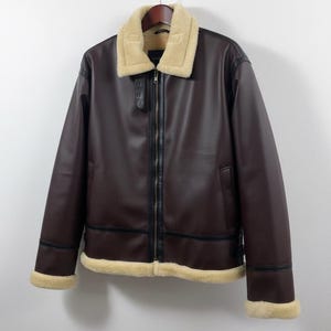 May include: A brown leather jacket with a cream-colored shearling collar and trim. The jacket has a zip-up front, black accents, and a classic aviator style. It hangs on a wooden hanger against a white wall.