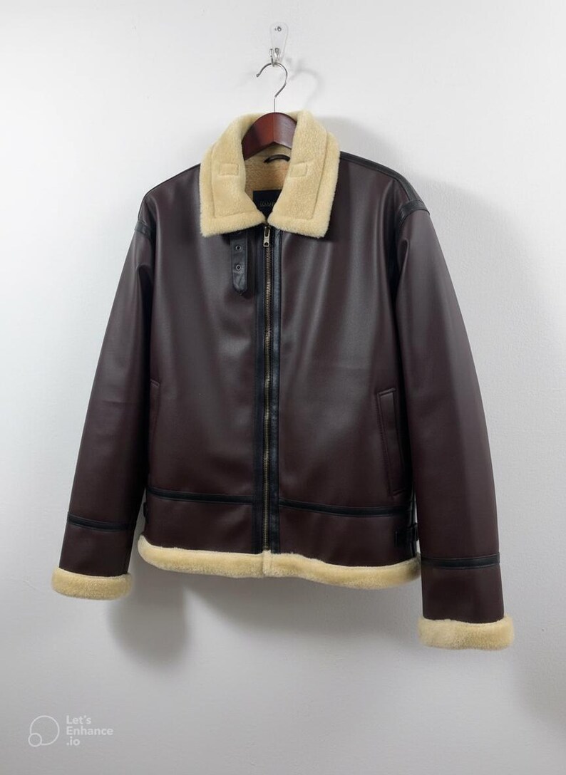 May include: A brown leather jacket with a cream-colored shearling collar and trim. The jacket has a zip-up front, black accents, and a classic aviator style. It hangs on a wooden hanger against a white wall.