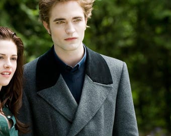May include: A gray double-breasted coat with a black collar. The coat is worn over a blue sweater and shirt. The background is a blurred green with a person on the left side of the image.