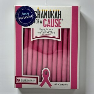 以下が含まれることがあります： ピンク色のハヌカのキャンドルの箱。箱には「CHANUKAH FOR A CAUSE」と「Happy Chanukah」の文字があります。ピンクのリボンのグラフィックと「45 Candles」の文字も表示されています。
