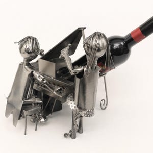 May include: A silver-toned metal wine bottle holder featuring two stylised figures crafted from wire and sheet metal. One figure sits at a miniature piano while the other stands beside it. The structure is designed to support a wine bottle horizontally.