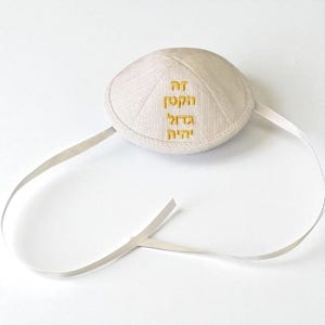 May include: Beige kippah with gold embroidered Hebrew text. The kippah is round with a stitched design and attached with a thin, off-white ribbon. The background is a plain white surface.