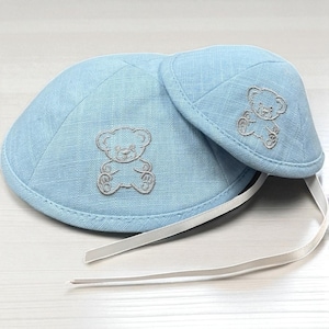 Puede incluir: Dos gorros de bebé azul claro con diseños de osos de peluche bordados. Los gorros están hechos de una tela texturizada y tienen un patrón acolchado. Una cinta blanca está unida a los gorros. Los gorros están diseñados para bebés.