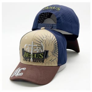 May include: A baseball cap featuring a tan front panel with a graphic of praying hands, a cross, and the text "JESUS LORD & SAVIOR". The brim is brown with a white "LC" logo. The back panels are navy blue with perforated holes and a snapback closure.