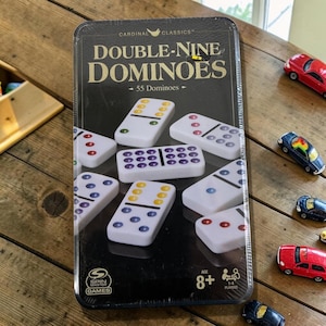 May include: A black rectangular tin of Cardinal Classics Double-Nine Dominoes, with 55 dominoes. The tin features the game title in large white letters. Dominoes are white with colored dots. The game is for ages 8+ and 1-4 players.