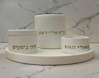 May include: A white ceramic set with gold Hebrew text, including a round tray, a cylindrical container, a small round container, and a candle. The set is designed for ritual use and features a minimalist aesthetic.