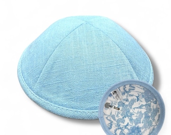 Könnte beinhalten: Eine hellblaue Kippa aus strukturiertem Stoff, neben einer passenden runden Anstecknadel mit floralem Design. Die Kippa ist eine traditionelle jüdische Kopfbedeckung, und die Anstecknadel scheint ein dekoratives Accessoire zu sein.