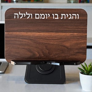 Puede incluir: Un letrero de madera marrón oscuro con el texto hebreo "Feliz día y noche" en letras blancas. El letrero tiene un patrón de vetas de madera y está montado sobre un soporte de metal negro. Una pequeña planta en maceta es visible en el fondo.