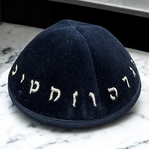 May include: Navy blue velvet kippah with embroidered Hebrew text. The kippah is a traditional Jewish head covering, featuring a rounded shape and a dark blue color. The embroidered text is in a lighter color.
