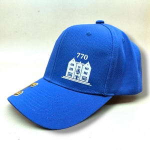 Könnte beinhalten: Königsblaue Baseballkappe mit gebogenem Schirm. Die Kappe zeigt eine weiße Grafik von drei Gebäuden mit der Nummer "770" darüber. Ein goldfarbenes Emblem befindet sich auf dem Schirm.