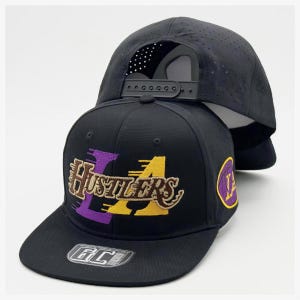 Pode incluir: Um boné snapback preto com painel traseiro perfurado e aba reta. A frente exibe o texto HUSTLERS bordado em marrom sobre letras grandes roxas e amarelas. Um emblema circular com um logotipo amarelo e roxo está localizado na lateral.